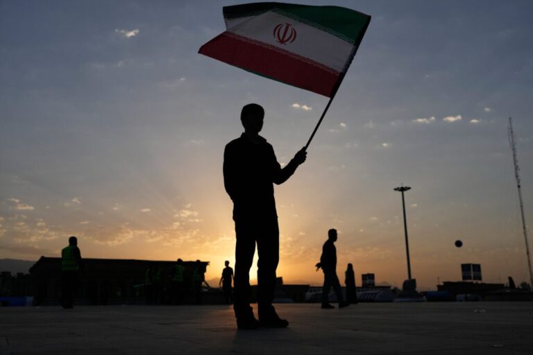 The sun rises as a man holds an Iranian flag on the esplanade of the Imam Khomeini Mosalla Grand Mosque before the start of the dayslong funeral ceremonies for slain Iranian Supreme Leader Ayatollah Ali Khamenei and members of his family in Tehran, Iran, Saturday, July 4, 2026. (AP Photo/Altaf Qadri)
